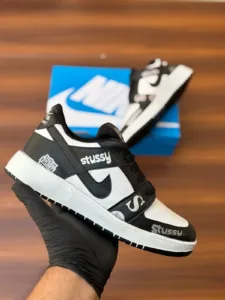 Budget-friendly streetwear sneakers with teal panels, cream swoop logo, yellow sole, and bold graphic patches, held in hand over a wooden background