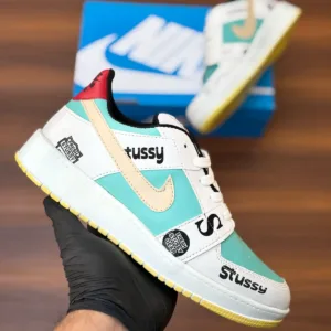 Budget-friendly streetwear sneakers with teal panels, cream swoop logo, yellow sole, and bold graphic patches, held in hand over a wooden background