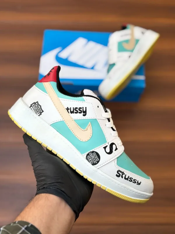 Budget-friendly streetwear sneakers with teal panels, cream swoop logo, yellow sole, and bold graphic patches, held in hand over a wooden background